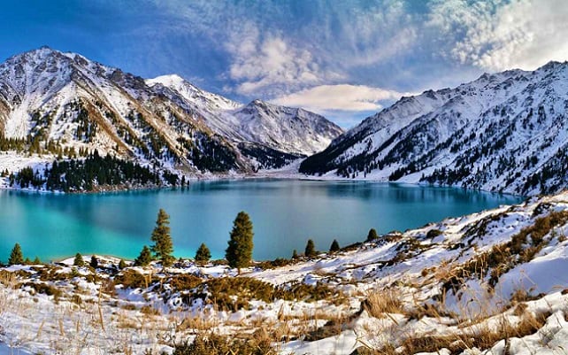 Turquoise mountain lake surrounded by snow-covered peaks and evergreen forests under blue sky with clouds in Kazakhstan